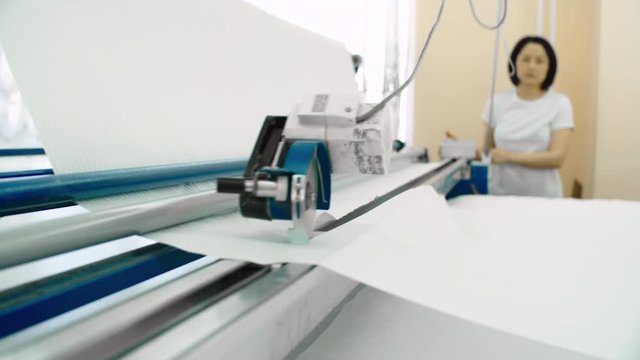 Rack focused shot of Asian female worker using textile cutting machine for cutting white fabric in factory