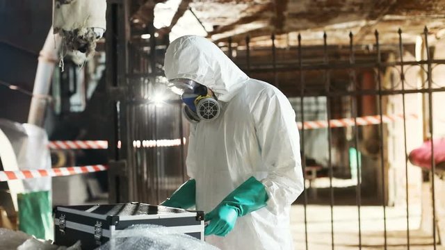 Young Caucasian man in white hazmat suit closing black suitcase, looking at camera. Two workers in colored hazmat outfits leaving old factory.