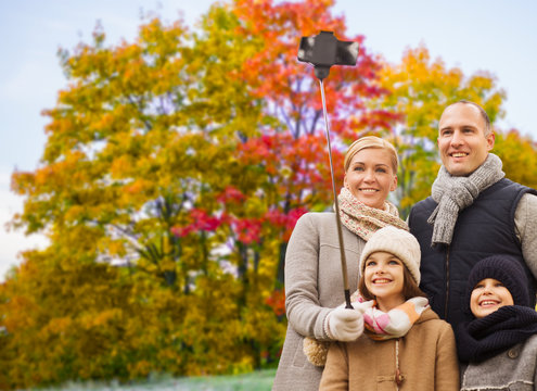 family, technology and people concept - happy mother, father, daughter and son taking picture by smartphone on selfie stick over autumn park background