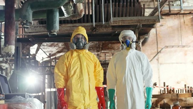 Slavic guys in yellow and white hazmat suits working stand in background of old factory after terrible accident. Two Caucasian scientists. Team. Indoors. Radiation.