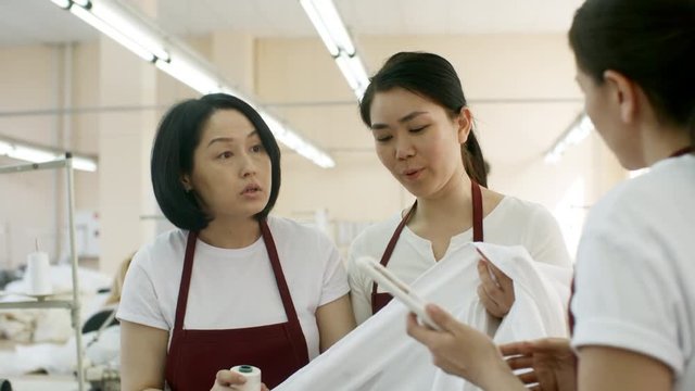 Asian seamstresses walking through sewing factory and discussing color of thread and fabric while female colleague showing something on digital tablet to them