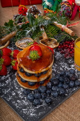 Christmas Breakfast Pancakes.  Stack of Pancakes with Blueberries and Strawberries drizzled with Maple Syrup, served on a slate platter with Christmas decorations, surrounded by Christmas gifts.