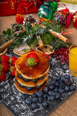 Christmas Breakfast Pancakes.  Stack of Pancakes with Blueberries and Strawberries drizzled with Maple Syrup, served on a slate platter with Christmas decorations, surrounded by Christmas gifts.