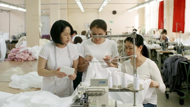 Panning shot of three asian women discussing white fabric and looking at digital tablet while working together in sewing factory