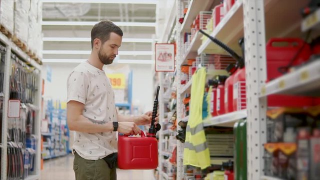 Male shopper is examining red plastic can for liquid with nozzle for car in store. Driver is choosing tools for garage