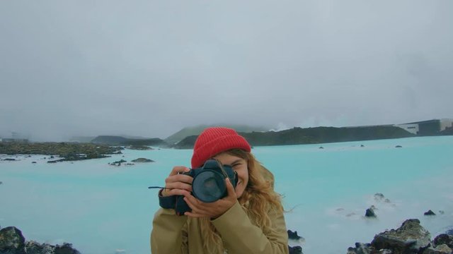 Young adventurous female photographer in brown rain coat and with red beanie walks around epic landscape of natural hot springs, blue silicon dioxide water. travel blogger destination
