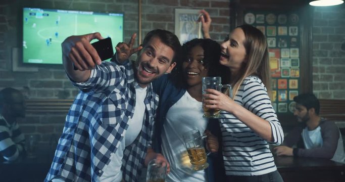 Young mixed races friends in the pub during sport game, man taking selfie photo on the smartphone with two pretty women.