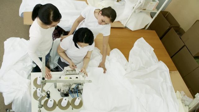 Top view of three seamstresses standing in textile factory, speaking and using overlock sewing machine while working with white fabric