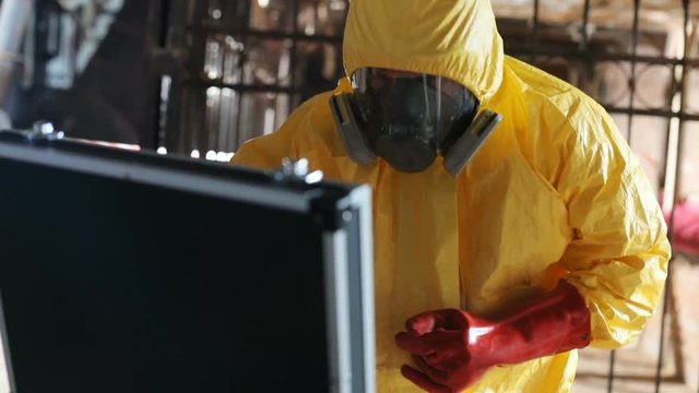 Guy in yellow hazmat outfit opening black briefcase, getting plastic bag with little stone. Indoors. Dangerous job. Biohazard.