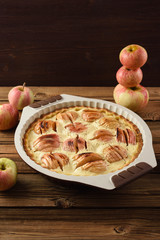 Homemade apple pie in baking dish served with raw organic apples on old wooden background