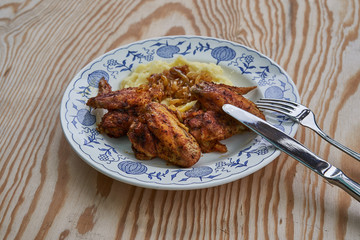 Close up Picture of old rustic plate with roasted spicy chicken wings, mashed potatoes with glazed, baked or fried onions and stainless steel fork, knife on wooden table.