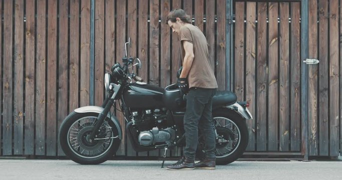 Handsome rider guy parked classic style motorcycle near wooden wall of garage. The biker inserts the key into the ignition lock and leaves from parking. Urban lifestyle 4k video