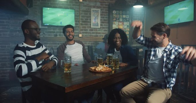Multiethnical friends sitting at the bar in the evening and giving five, cheering up while watching sport game.
