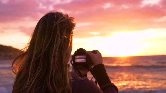 CLOSE UP, LENS FLARE: Unknown woman taking photos of the ocean and the beautiful summer sunset on windy evening. Unrecognizable girl on summer holiday capturing the waves and sunrise with her camera.