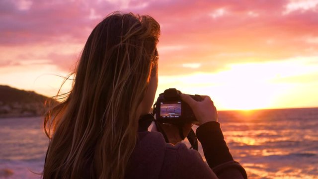 SLOW MOTION, CLOSE UP: Young female photographer capturing the picturesque summer evening sun above the ocean. Gentle breeze blows through girls hair while she takes photos of the scenic sunset.