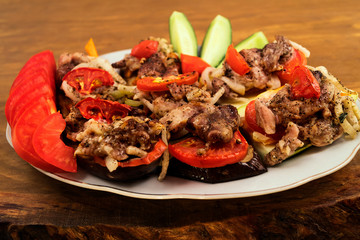 A portion of fried meat with baked eggplant, zucchini, tomatoes and onions, garnish of sliced fresh cucumbers and tomatoes, wooden background.