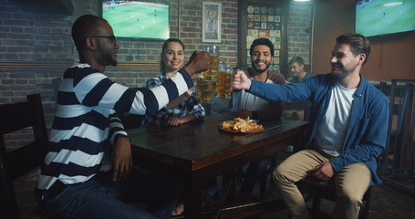 Multiethnical team of male and female sport fans doing cheers with beer while watching sport game at the bar.