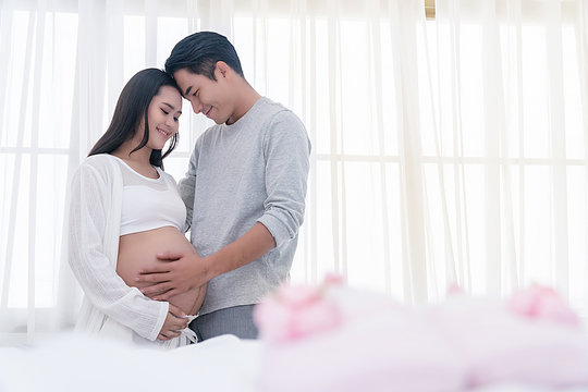 Husband embracing his wife with love beside the white window. Happy couple of young man hugs his pregnant beautiful wife, smiling and looking into each other's eyes at home. Concept of expecting baby.