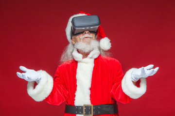 Christmas. Santa Claus in black virtual reality glasses makes gestures with his hands. Surprise, emotion. New technology. Isolated on red background.