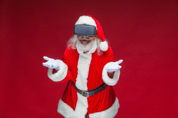 Christmas. Santa Claus in black virtual reality glasses makes gestures with his hands. Surprise, emotion. New technology. Isolated on red background.