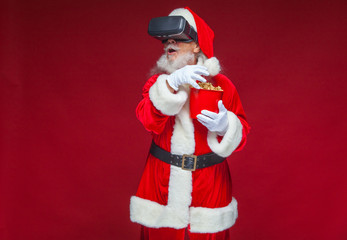 Christmas. Santa Claus in black virtual reality glasses holding a red bucket of popcorn. Watching a movie in virtual reality. Isolated on red background.