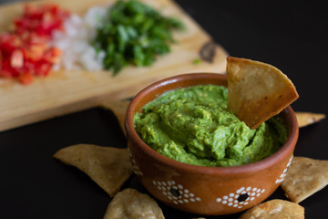 Guacamole mexicano tradicional de la salsa en el cuenco de arcilla y nachos en el fondo de un tablero de pizarra