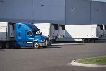 Big rig semi truck with refrigerated semi trailer standing for loading cargo in warehouse dock with another refrigerator semi trailers