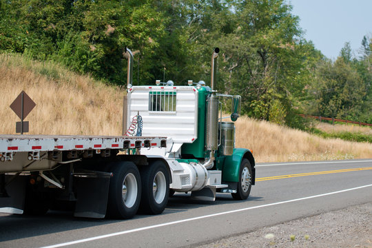 Green big rig day cab semi truck with empty flat bed semi trailer driving to point of loads