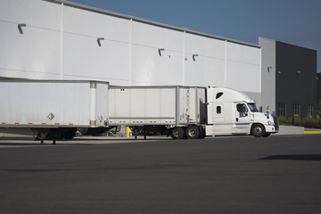 White big rig semi truck with dry van semi trailer stand in warehouse dock with another semi truck...