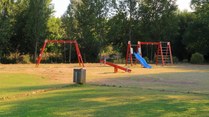Parque para ni&ntilde;os en paisaje verde con columpios de madera de color rojo y arboles en el fondo en verano al atardecer