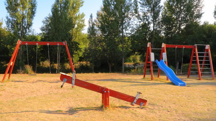Fototapeta premium Paisaje con parque para niños con columpios de madera de color rojo y fondo de arboles y cielo azul al atardecer