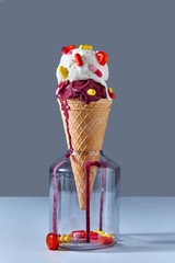The waffle cone with a cold ice cream stands in glass jar with red and yellow capsules and a lollipop. Jar on a gray background. Pils and lollipops in a jar.