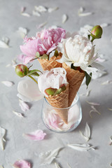 Greeting card with three sweet wafer cones and gentle peony flowers in a glass vase, petals on a gray concrete background.