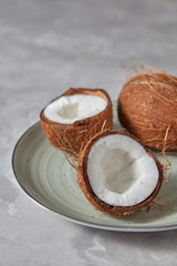 The halves of tropical coconut with whole coconut on a plate on a gray.