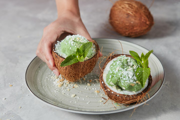 Womans hands take a half of coconut with homemade green ice cream from plate on a gray concrete.