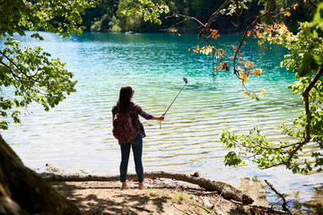 Back view of tourist woman with backpack standing on lake shore taking picture. Beautiful colorful spring panorama of lit by sun green forest and blue lake on background