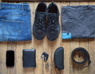 Men's casual outfits. Men shoes, clothing and accessories on wooden background - grey t-shirt, blue jeans, sneakers with eyeglasses, wallet, belt. Top view. Flat lay.