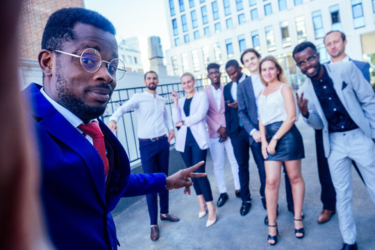 successful and beautiful American afro man in a business suit in a white shirt looking into the phone and takes photos selfie standing company of people on the street . group of nine businesspeople
