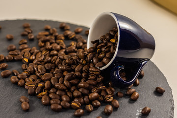 Eine Espresso-Tasse gefüllt mit ungemahlenen Kaffeebohnen liegt auf einer schwarzen Platte. Die Kaffeebohnen fallen aus der Tasse und weitere ganze Kaffeebohnen liegen herum.