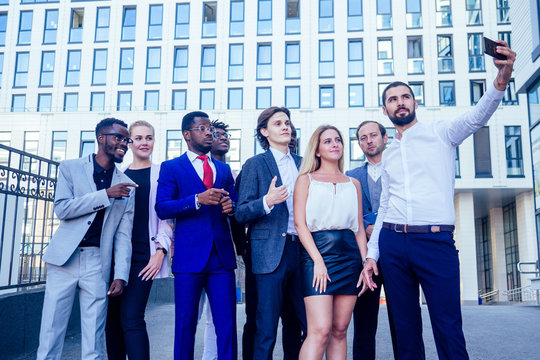 successful group of nine multinational businesspeople in a business suit looking into the phone and takes photos selfie company of people on the street against the background of a skyscraper window.