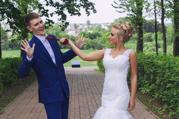 Funny wedding couple outdoors. Bride holding groom for tie. Cheerful marriage concept.
