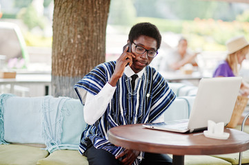 African man in traditional clothes and glasses sitting behind laptop at outdoor caffe and speaking on mobile phone.