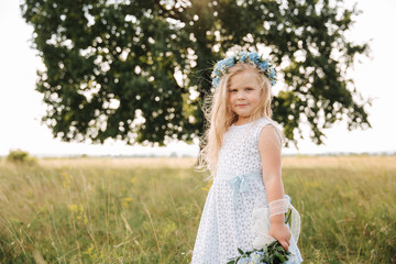 Fototapeta na wymiar Little girl in sky blue dress with wreath and bouquet stand in field in front of big tree