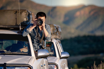Young Asian male traveler and photographer sitting on the car window taking photo on road trip in...