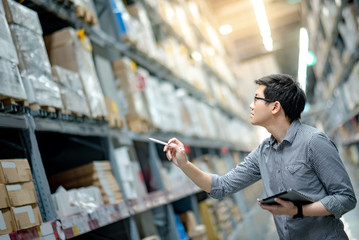 Young Asian man worker doing stocktaking of product in cardboard box on shelves in warehouse by using digital tablet and pen. Physical inventory count concept - obrazy, fototapety, plakaty