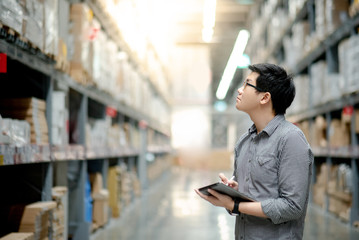 Young Asian man worker doing stocktaking of product in cardboard box on shelves in warehouse by using digital tablet and pen. Physical inventory count concept