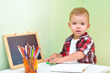 Two year old baby boy in red checkered shirt sitting at table with an album and colored pencils