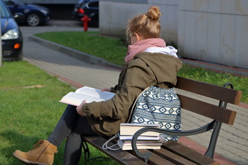 Młoda blondynka uczesna w kok, w zielonej kurtce, różowym szaliku, jeansach i trzewikach, siedzi na ławce na osiedlu mieszkaniowym czyta książkę, obok niej leży kilka innych książek, plecak