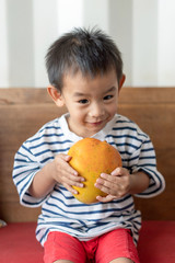 Fototapeta premium adorable little boy holding fresh ripped mango. Asian little boy sitting on wooden sofa with fresh huge mango in his hands and smile on his face with lovely atmosphere.
