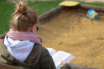 Nastolatka (dziewczynka, młoda kobieta) siedzi na skraju piaskownicy i czyta grubą książkę, blond włosy ma upięte w kok, różowy szalik, zieloną kurtkę, okulary słoneczne, widok z boku - obrazy, fototapety, plakaty